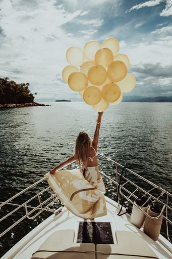 A vertical shot of a woman in a dress standing on a yacht, holding golden balloons while sailing