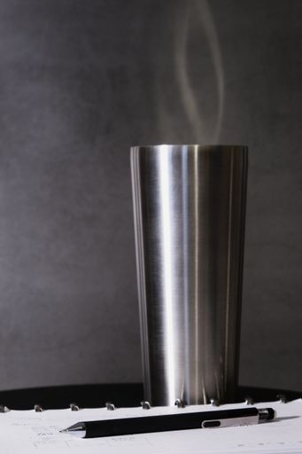 A vertical shot of a hot liquid in a big metallic cup on the table near a notepad with a pen