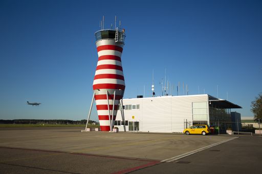 Horizontal view of Red and white control tower of Lelystad Airport against a clear blue sky. View of terminal with small plane flying by.