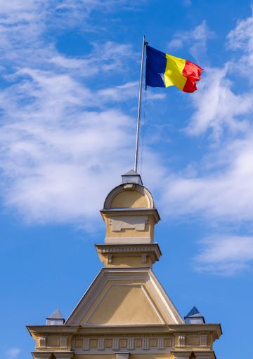 A vertical shot of a Romanian flag on top of a building against a blue cloudy sky