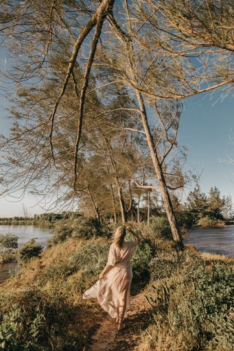 A vertical shot of a woman in white dress walking barefoot in small grassy area surrounded by water