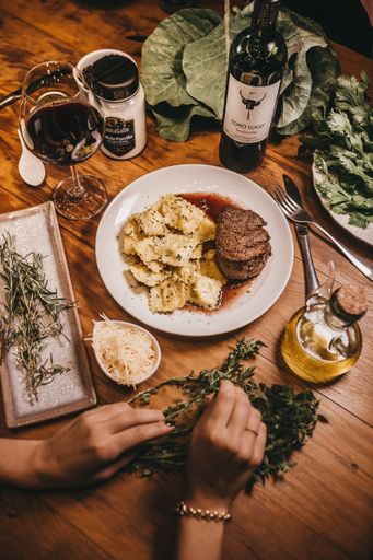A vertical closeup shot of a plate with ravioli and steak next to a wine bottle and a wine glass