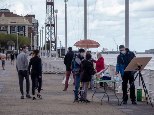 A photo of a man painting a picture on the riverbank while passersby are looking at his drawing