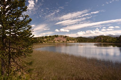 A landscape view of a lake surrounded by mountains under a blue cloudy sky on a sunny day