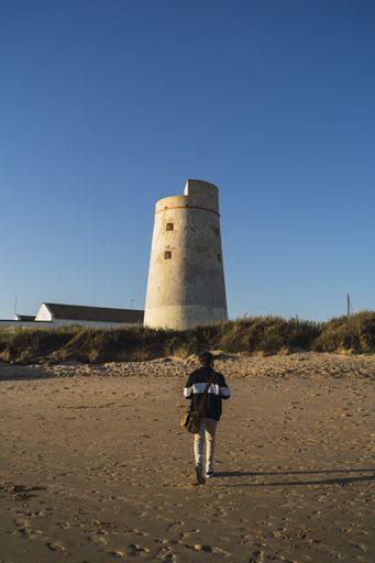 A young man with a bag walking towards a cylindrical tower in the beach