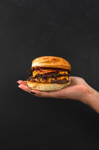 A vertical shot of a hand holding a juicy burger with cheese and bacon on a black background