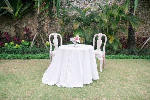 A wide shot of a small round table with a white table clothing and two chairs with the background of green plants