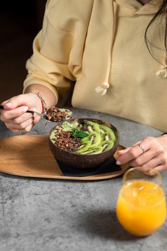 A vertical shot of female hands eating tasty oatmeal with kiwi and chocolate on the table with a cup of orange juice