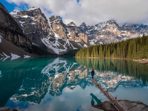A panoramic view of a girl standing on the edge of a tree trunk over Moraine Lake surrounded by mountains and trees reflecting on the water