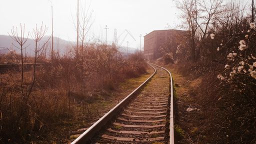 A train track between dry plants and trees with a building and clear blue sky in the background