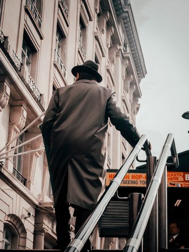 A vertical back view of a man with a hat and a vintage coat walking up the stairs in London, UK