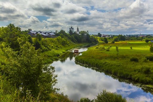 An aerial shot of a boat sailing on a river in the middle of a grassy field with trees under a cloudy sky