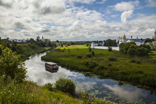 An aerial shot of a boat sailing on a river in the middle of grassy field under a cloudy sky