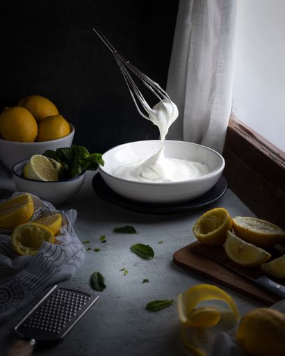 A vertical shot of a whisk in the air making meringue surrounded by fresh cut lemon and mint leaves.