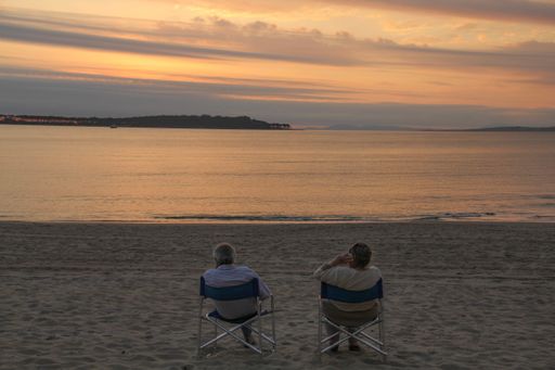 Two old people sitting on the folding chairs at a beach surrounded by the sea during the sunset