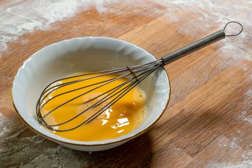 A white bowl of beaten egg with a whisk put on the table covered with flour