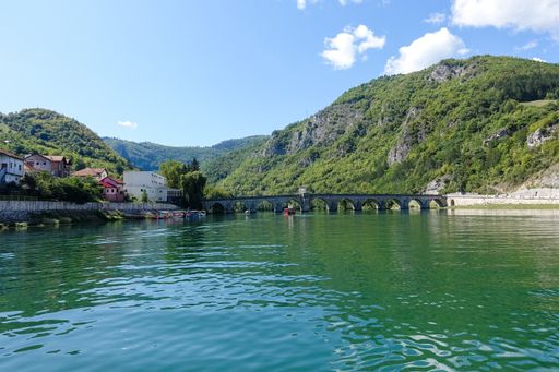 A scenic view of green lake water with houses on the shore, bridge, and mountain ranges on background