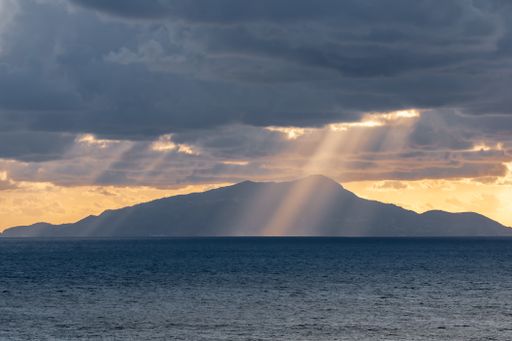A beautiful shot of sunlight peeking from behind the clouds illuminating the silhouette of Mount Vesuvius behind the sea, Italy