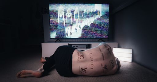 A closeup of a Man covered in branded tattoos laying in front of the TV showing a catwalk