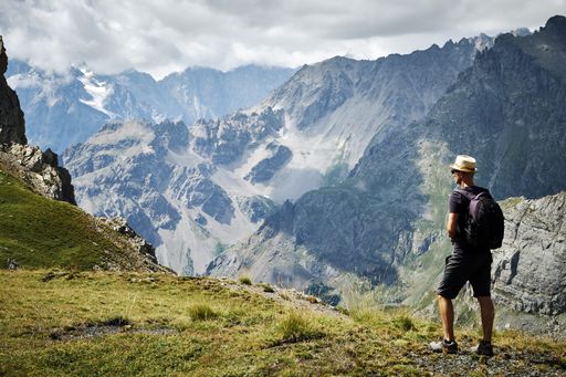 A landscape shot of beautiful mountains, a man with a backpack and a hat standing in the green field. Great for a hiking piece.