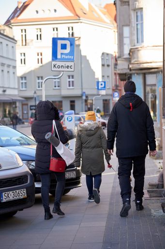 A back view of people walking on a sidewalk with parked cars on Ostrowek street