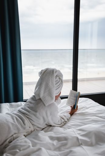 A lonely female with a white robe and a towel lying down in the bed and reading a book near the window