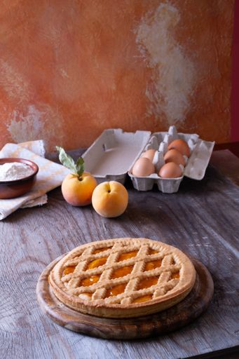 A vertical shot of an apricot pie with apricots, fresh eggs and a bowl of flour on a table