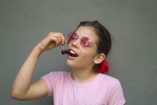 A young girl wearing sunglasses while eating cherries isolated on a gray background