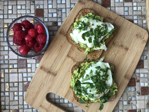 A vertical top view of two avocado toasts on a cutting board, and a small bowl of raspberries