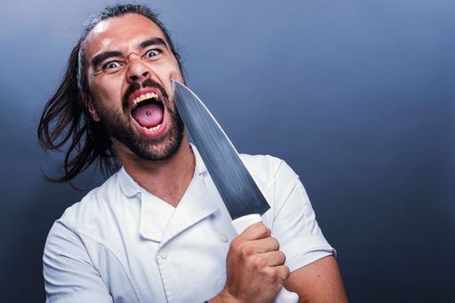 A closeup shot of a screaming male holding a knife and standing in front of a gray wall