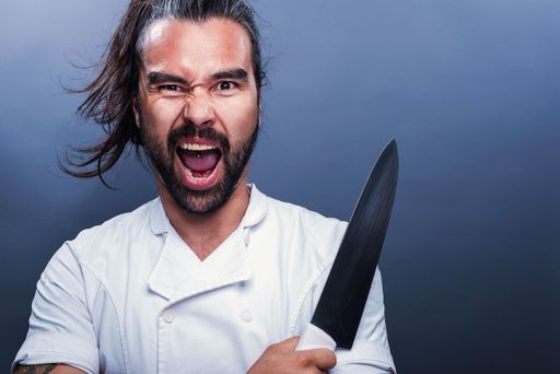 A closeup shot of a screaming male holding a knife and standing in front of a gray wall