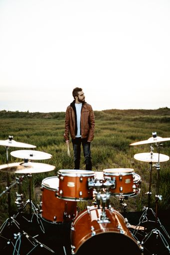A vertical shot of a guy with drums sticks standing behind a brown drum set in the middle of a field