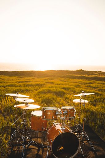 A vertical shot of a brown drum set on a black mat in the middle of a green meadow on a bright day