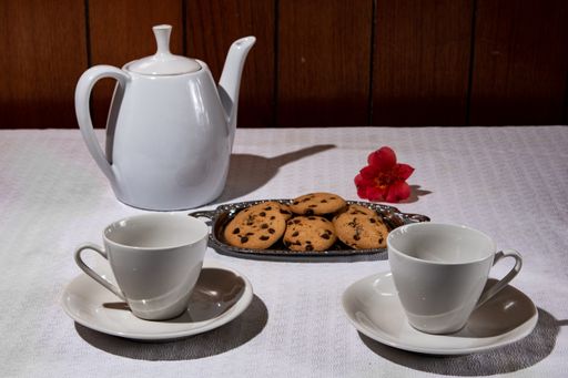 A closeup of a ceramic teapot with cups and chocolate chip cookies on the table under the lights