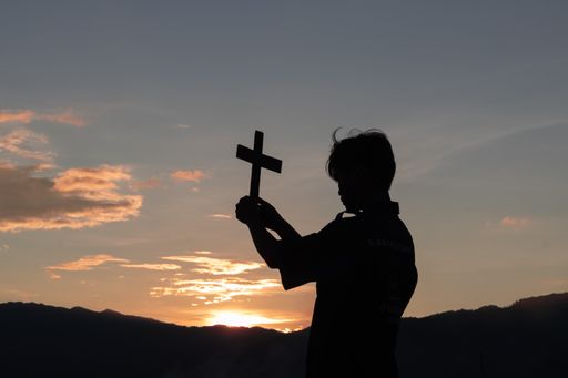 A Silhouette of a male holding Christian cross in hands to praying for blessing from god at sunset background