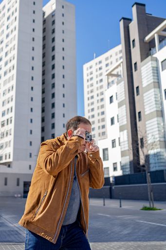 A closeup of an older man photographing with an old film camera in the city with buildings in the background