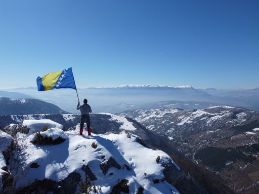 A man, with his back turned, standing on a mountaintop and waving the flag of Bosnia and Herzegovina