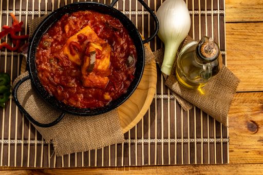 A high angle closeup shot of a pot of red soup with fish next to onion and oil on a wooden table