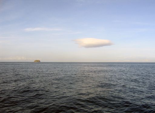 A wide shot of a small islet on the body of the sea in the distance under a clear blue sky