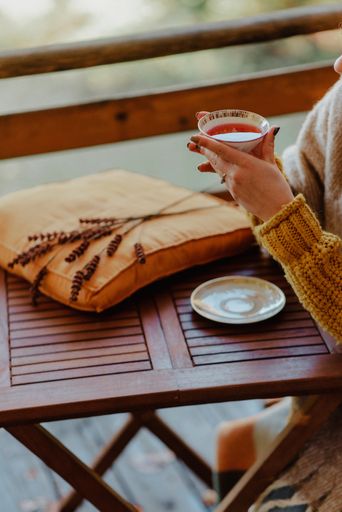 A vertical shot of a Caucasian woman's hand holding a cup of tea, near a wooden table with pillow and decorative plant on it