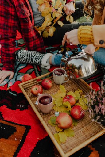 A closeup shot of two cups of tea being poured from a teapot with apples, leaves and flowers beside, during a picnic