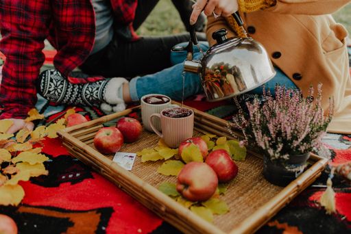 A closeup shot of two cups of tea being poured by a teapot, with apples, leaves and flowers beside, during a picnic