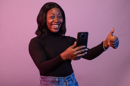 An excited Nigerian woman holding her phone with thumbs up isolated on pink background