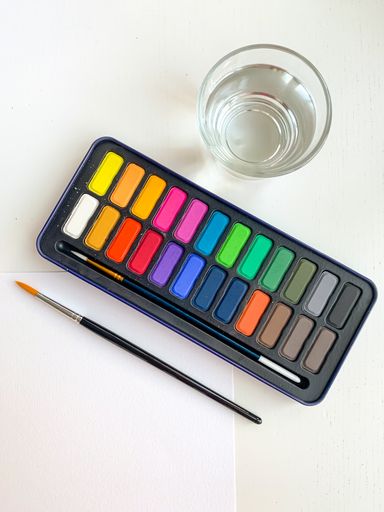 A vertical top view of new watercolors and brushes, white paper, and a glass of water