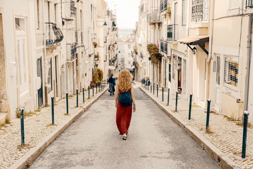 A young female tourist in a red dress walking through a road surrounded by buildings under the sunlight
