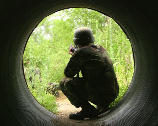 A United States military man with a rifle squatting down in a drainage pipe.