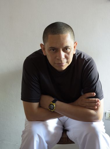 A vertical portrait of a Hispanic male sitting with crossed arms