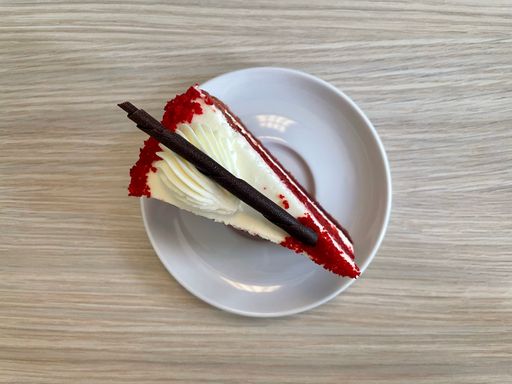 A top view of a piece of red velvet cake decorated with roll made of  chocolate on a white plate lying on a wooden table.