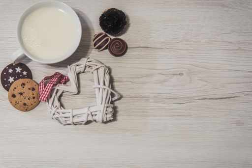 An overhead top view of a cup of fresh milk with various candies and cookies on a wooden surface
