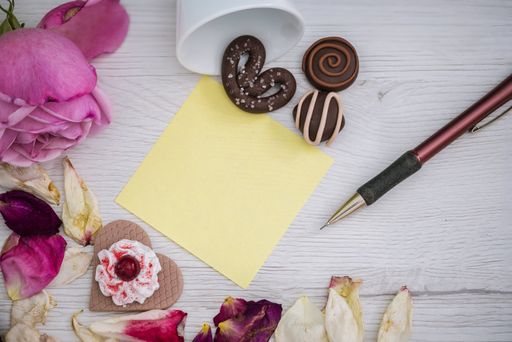 An overhead top view of a pink rose, petals, chocolate candies and cookies around a notepad on a wooden background